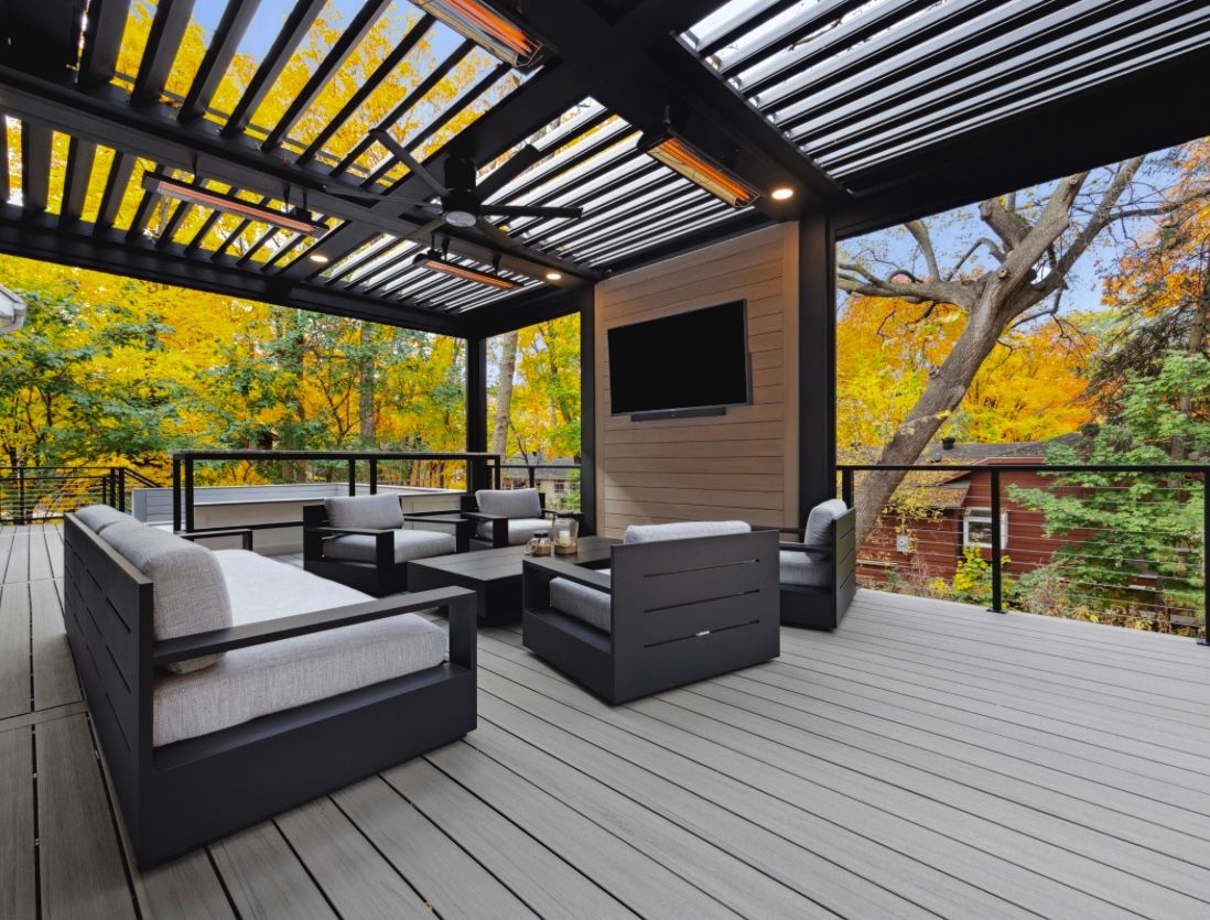 Outdoor deck with modern furniture, pergola, and TV, surrounded by fall foliage.