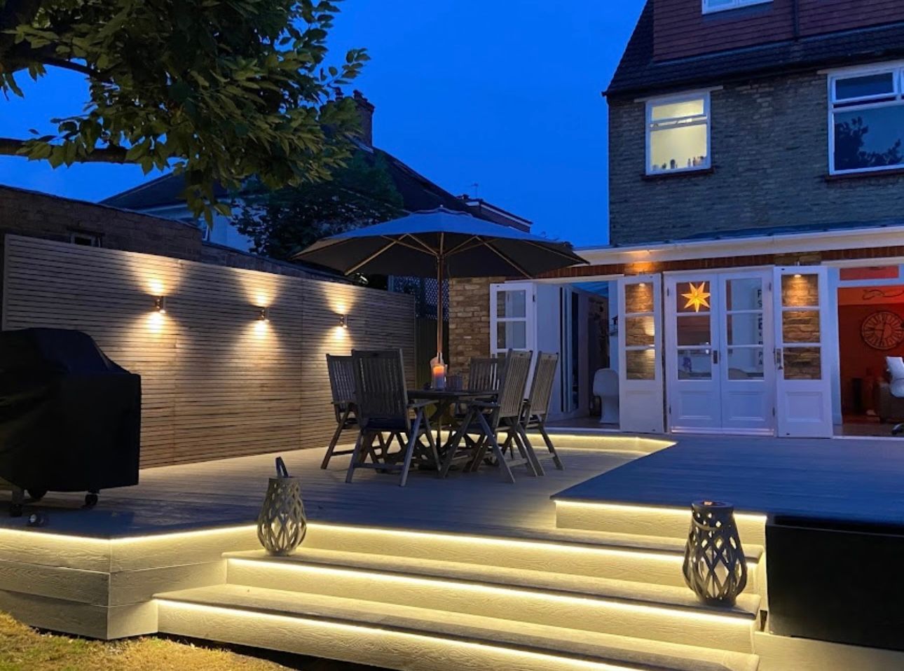 Outdoor patio at night with ambient lighting, a table, chairs, and a grill, near a house.