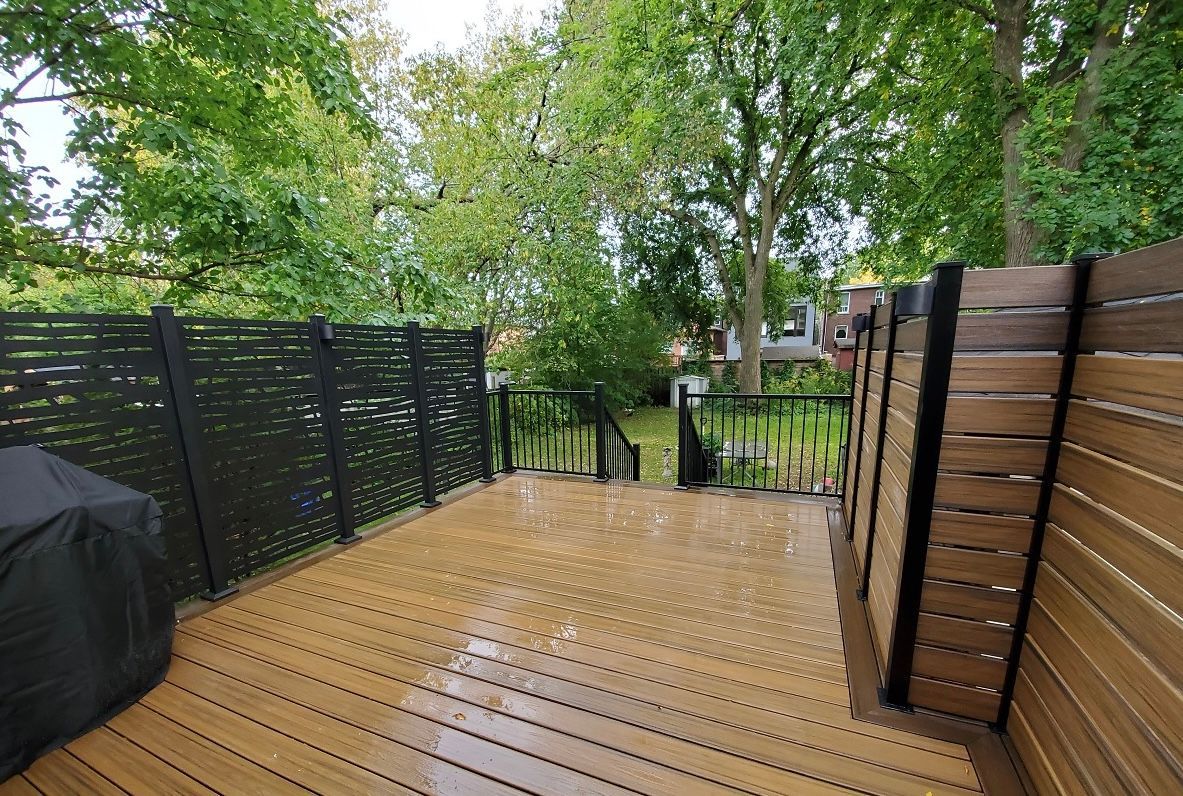 Wooden deck with black and wood-tone fences, leading to a grassy area with trees; overcast day.