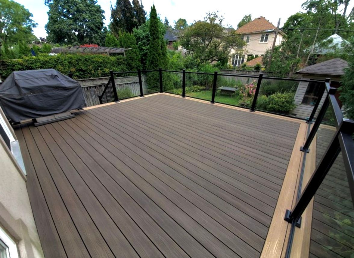 A brown composite deck with black railing and glass panels, overlooking a residential backyard.