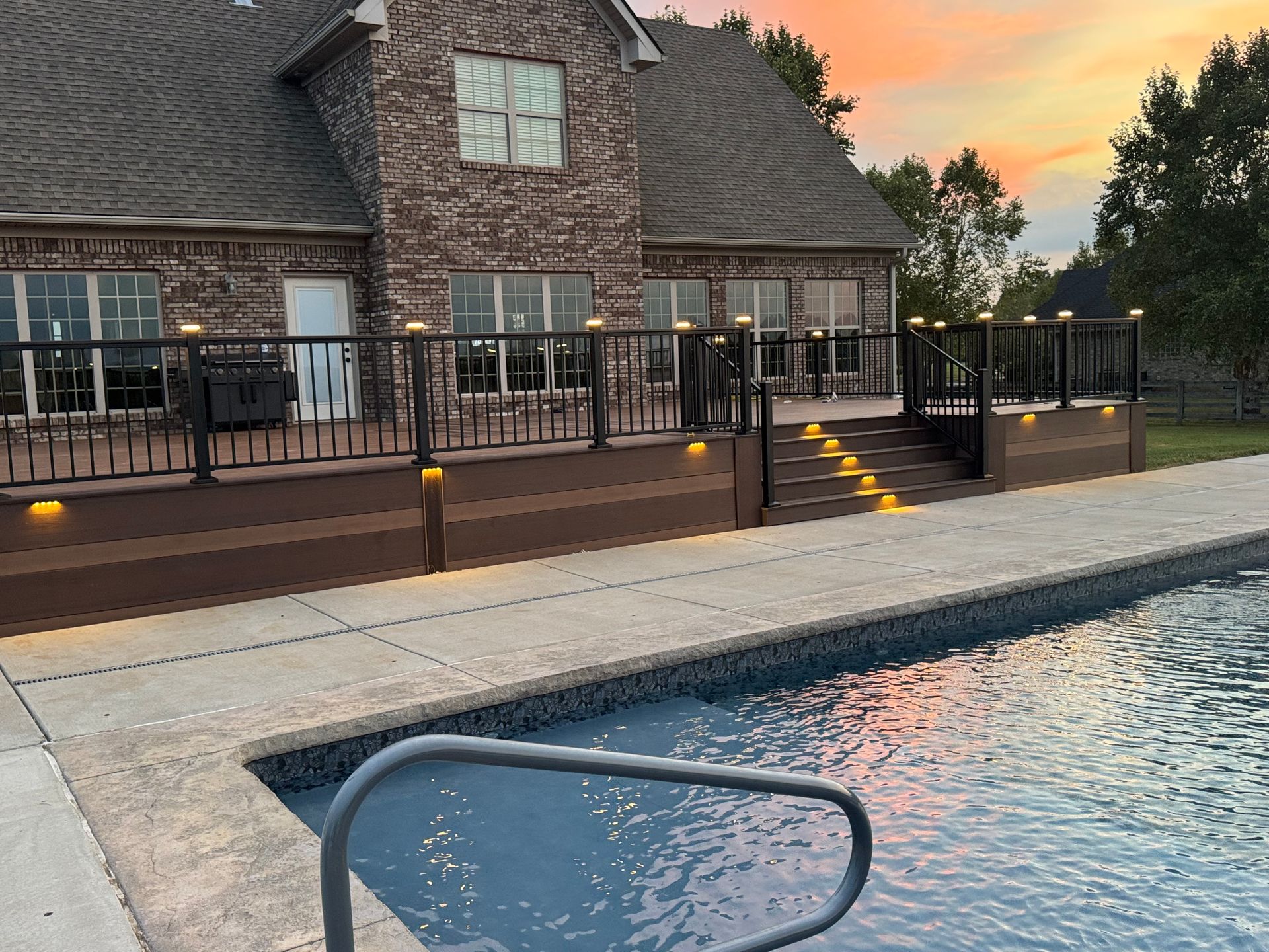 A backyard with a pool, deck, and house at sunset. Brown deck with black railing and lights.