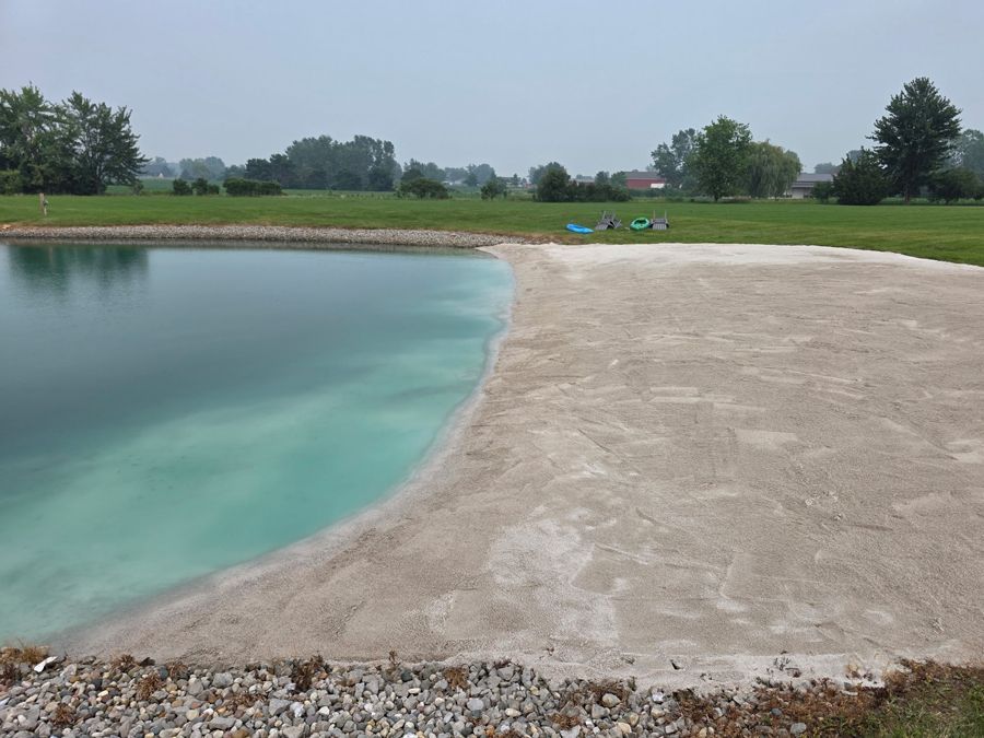 A turquoise pond beside a sandy shore in a grassy park under an overcast sky