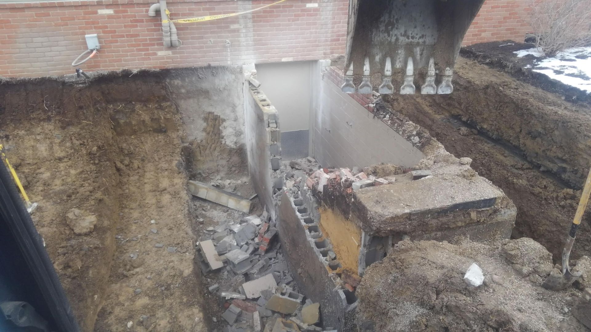 Collapsed excavation beside brick buildings, with rubble, exposed soil, and a damaged wall or trench.