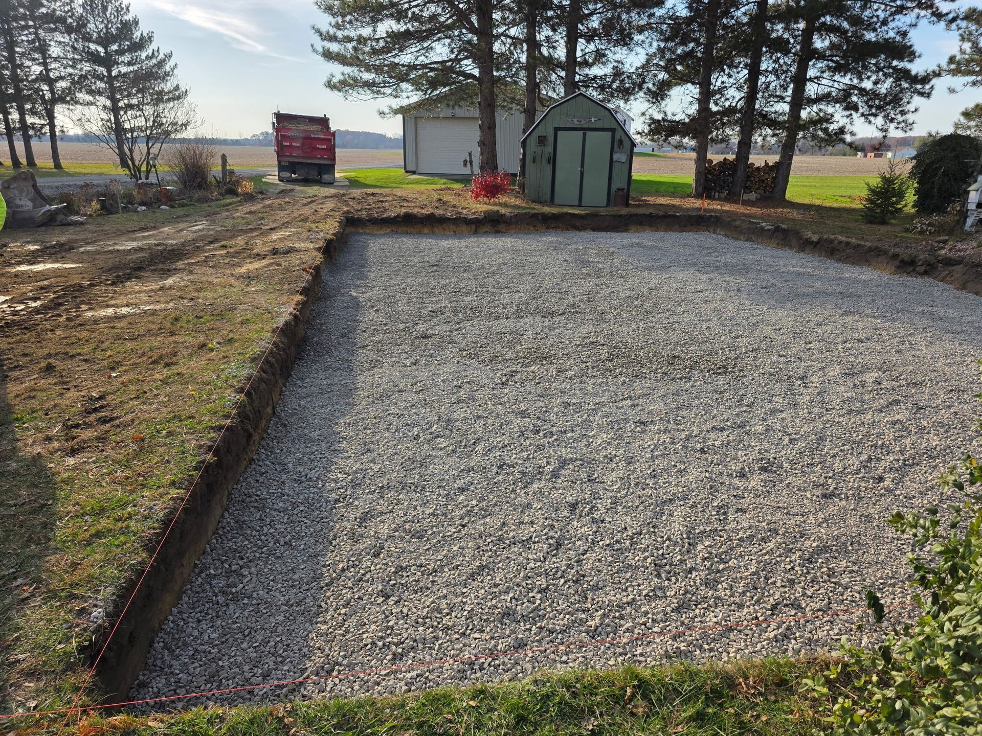 Gravel-filled rectangular area, bordered by grass, likely for a structure. Small red truck, shed, and trees in background.