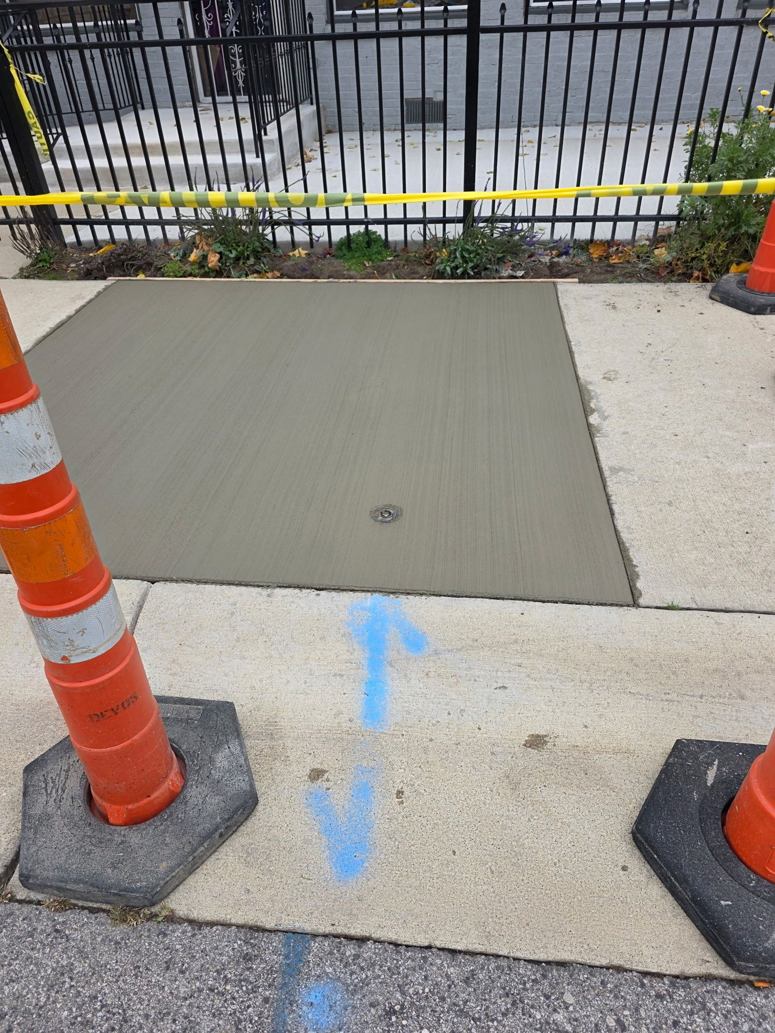 Fresh concrete patch on a sidewalk with an arrow, orange cones, and caution tape.