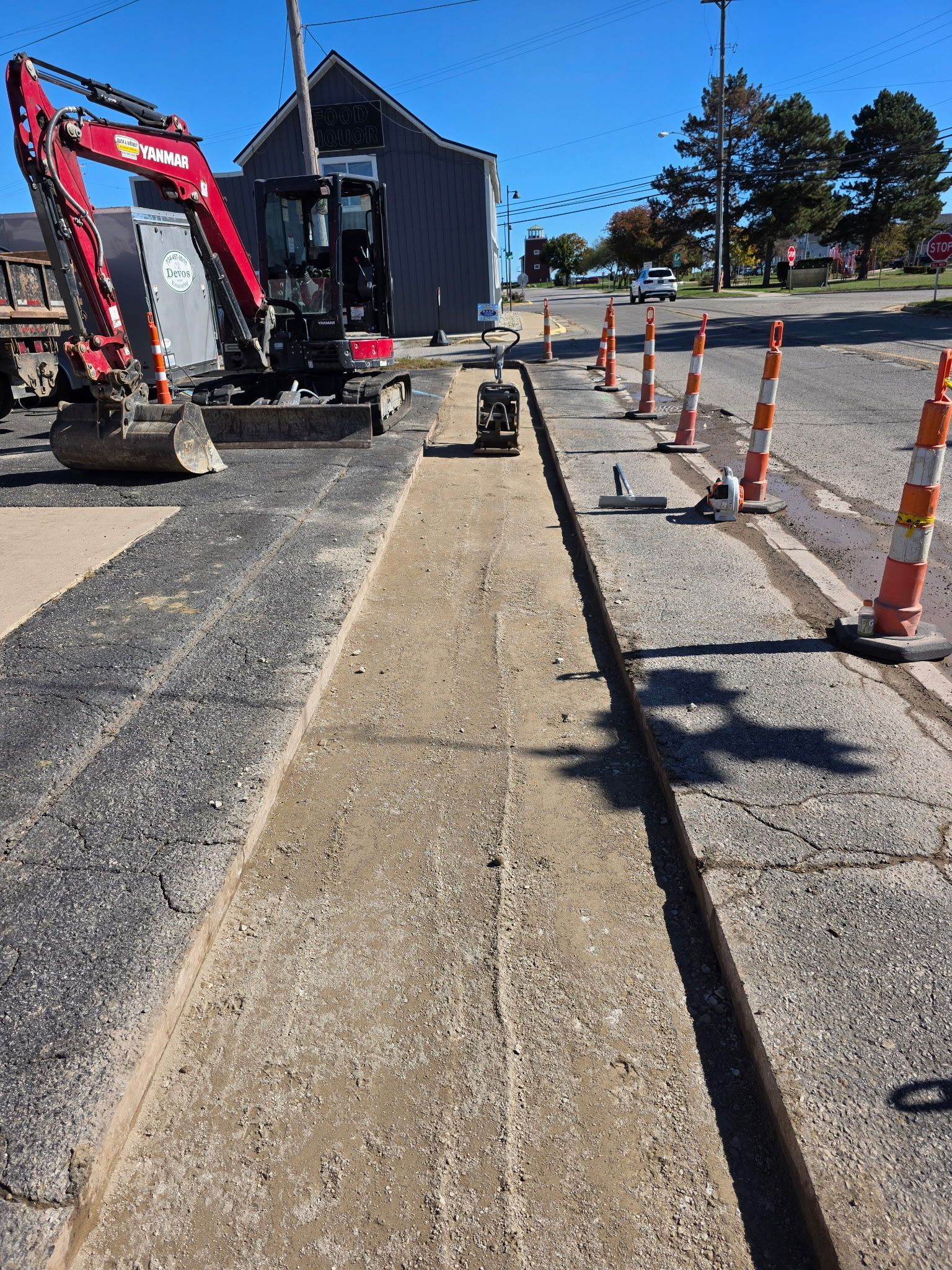 Road construction: excavator, trench, orange cones, building in background, sunny day.