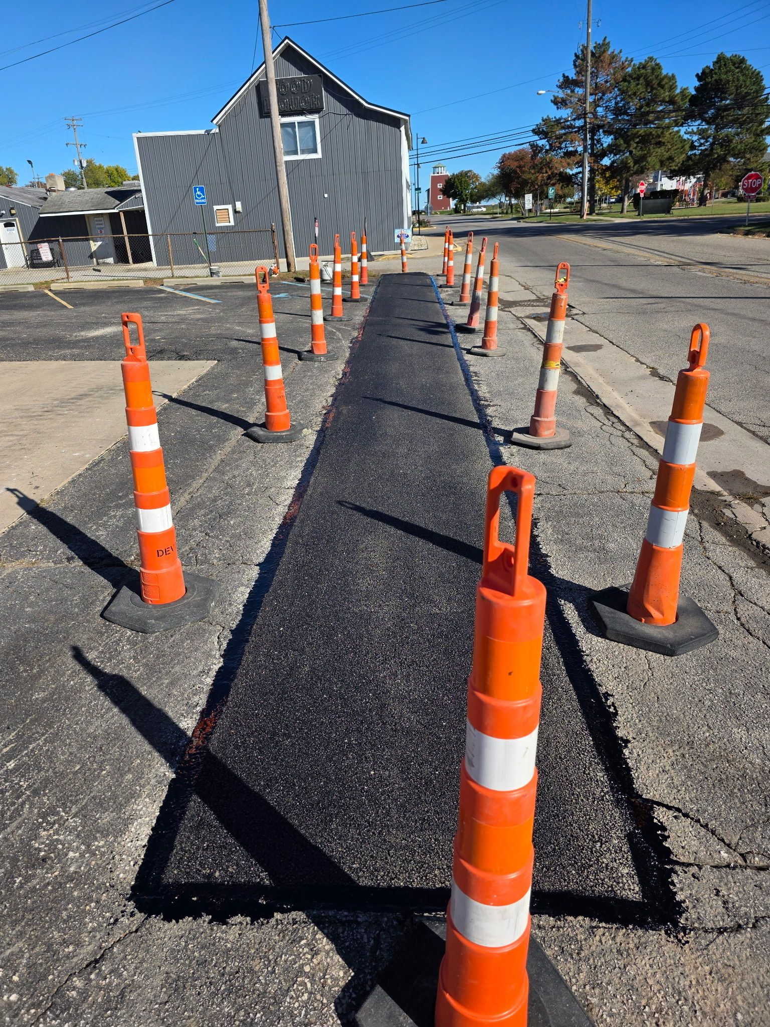 Road repair: Freshly paved strip of asphalt, marked by orange cones, in a street. Building in background.