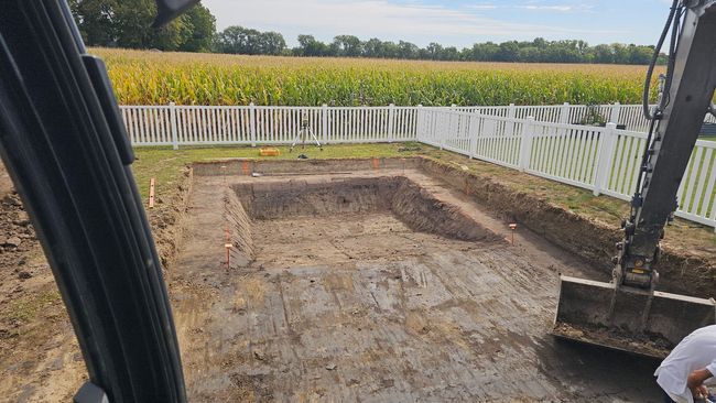 Excavated rectangular hole in ground, bordered by white fence. Field of corn in background.