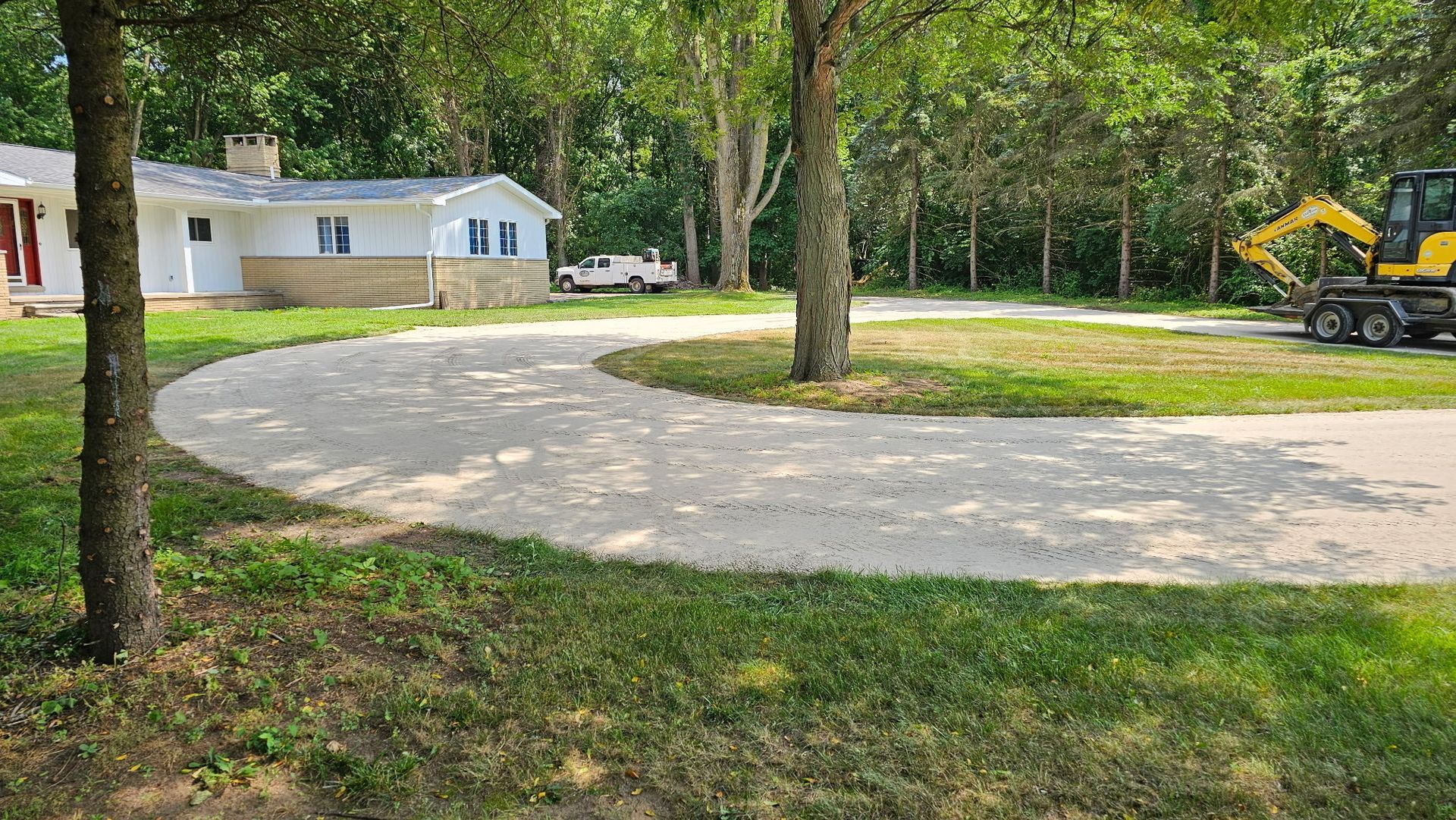 Gravel driveway curves past a white house and trees. An excavator and truck are parked in the distance.