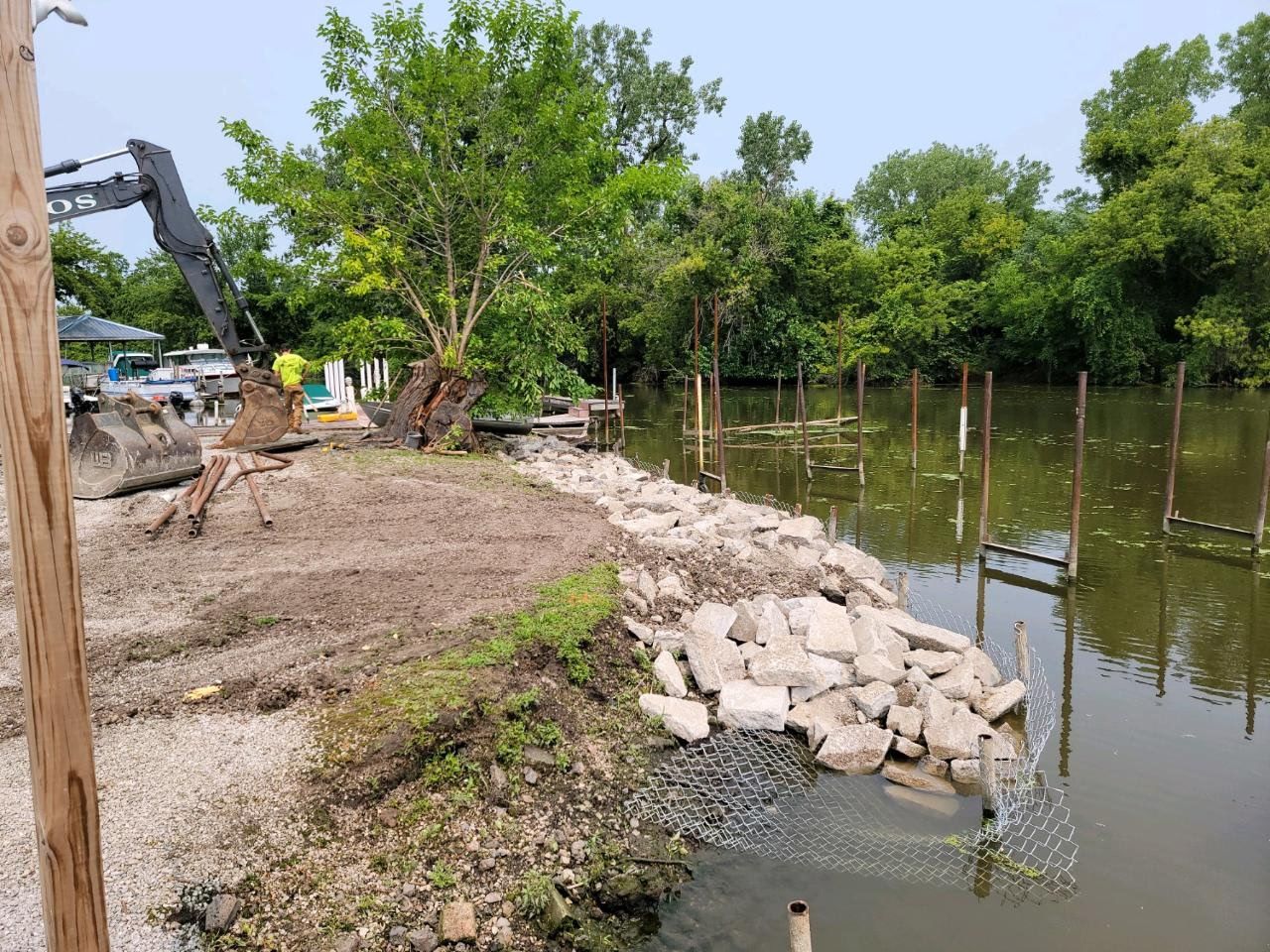 Shoreline with rocks, muddy bank, and a small canal beside dense green trees and shrubs