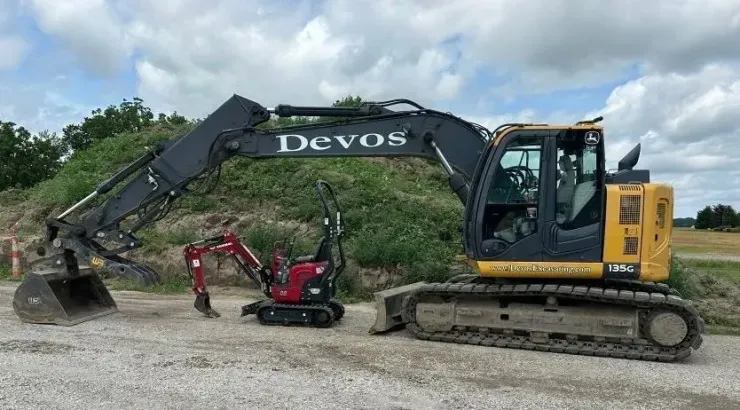 Yellow excavator with Devos boom arm lifting debris near a small red machine on a gravel lot under cloudy skies