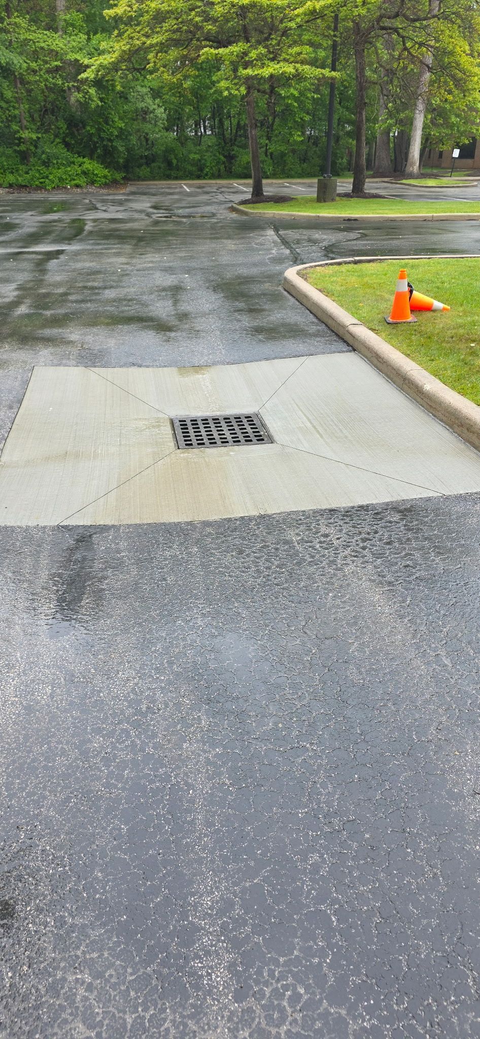Gravel road beside a wooded area, with a concrete curb, drain grate, and orange cone on the right.