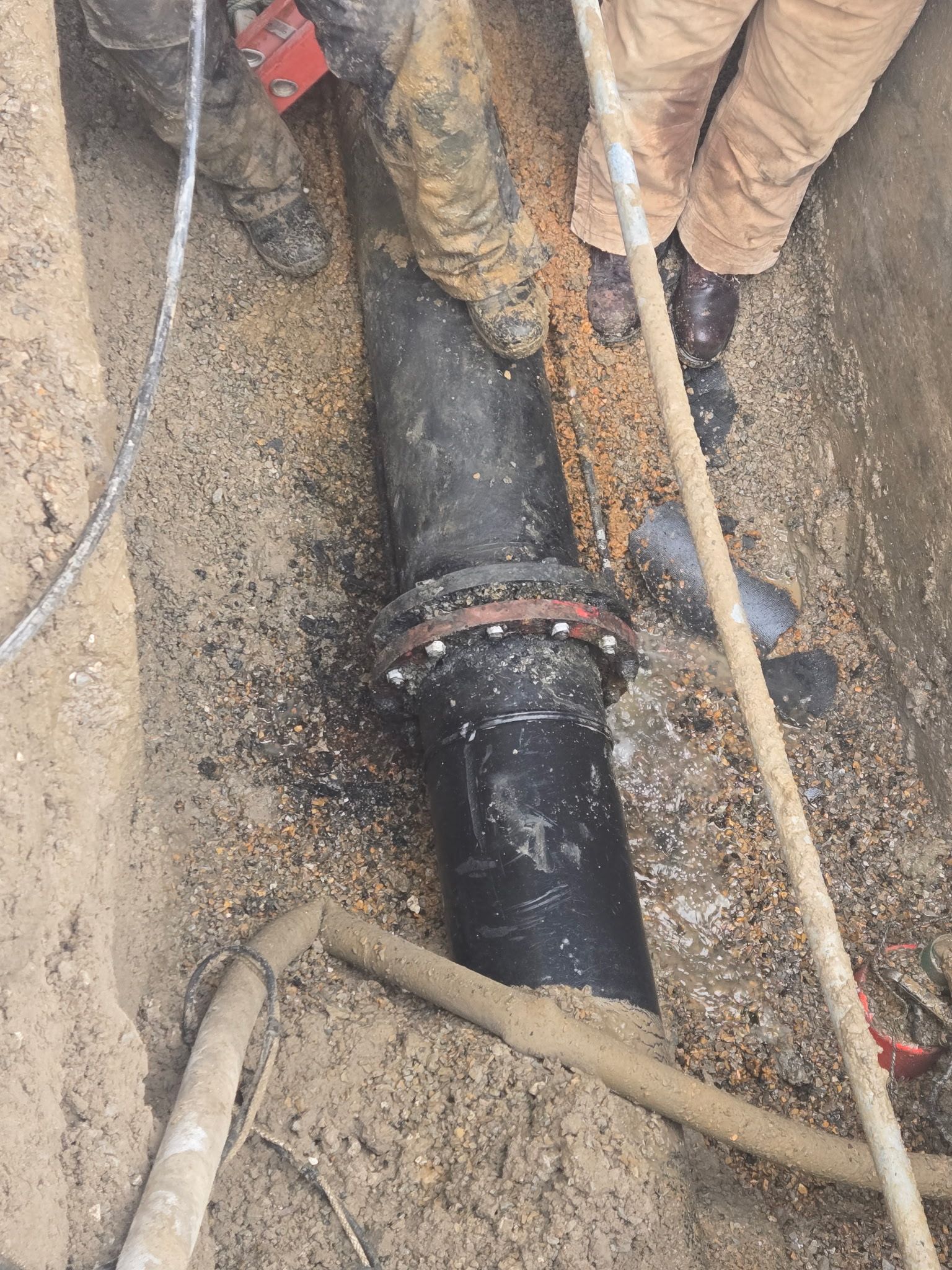 Workers repairing a black underground pipe in a muddy trench with tools around it