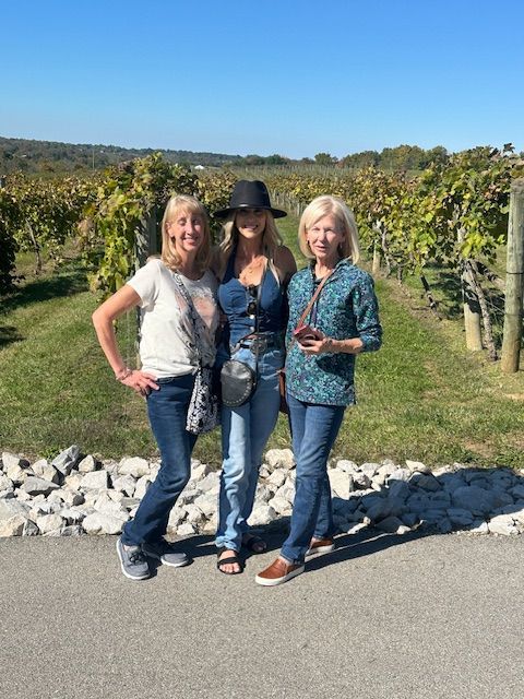 Three women are posing for a picture in front of a vineyard.