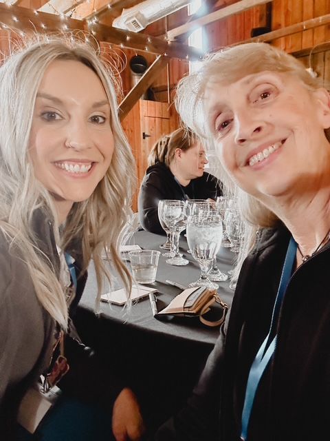 Two women are posing for a picture while sitting at a table with wine glasses.