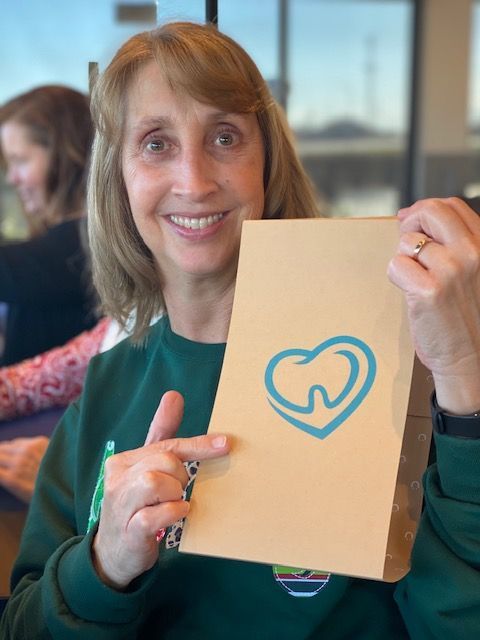Woman smiles, pointing at a brown paper bag with a heart-shaped logo containing a tooth.