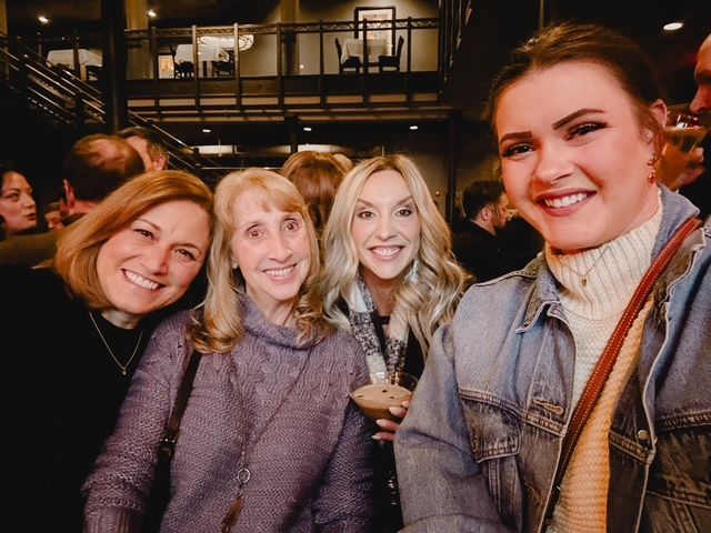Four smiling women take a selfie in a crowded venue.