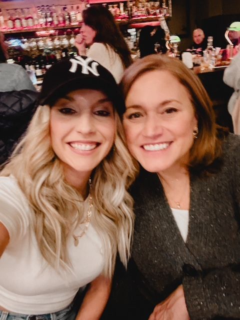 Two women smiling, posing for a selfie in a bar setting.