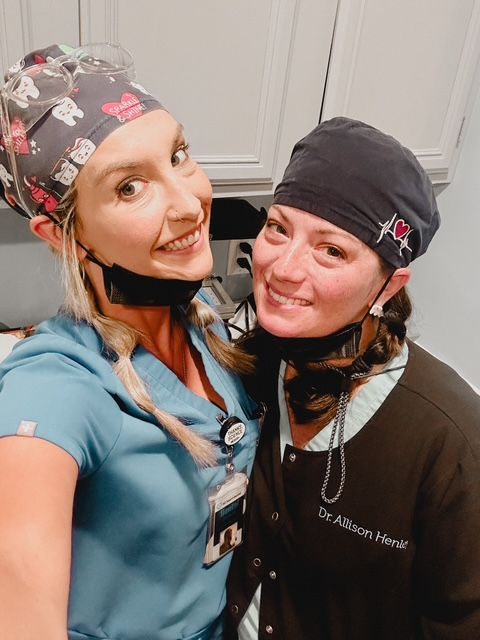 Two smiling dental professionals in scrubs and caps, one blonde, one brunette, indoors.