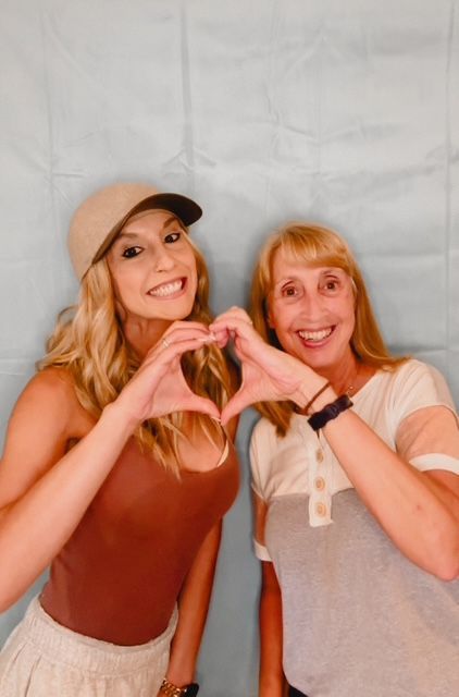Two women, one in a cap and brown top, the other in a gray and white shirt, making a heart shape with their hands, smiling at the camera.