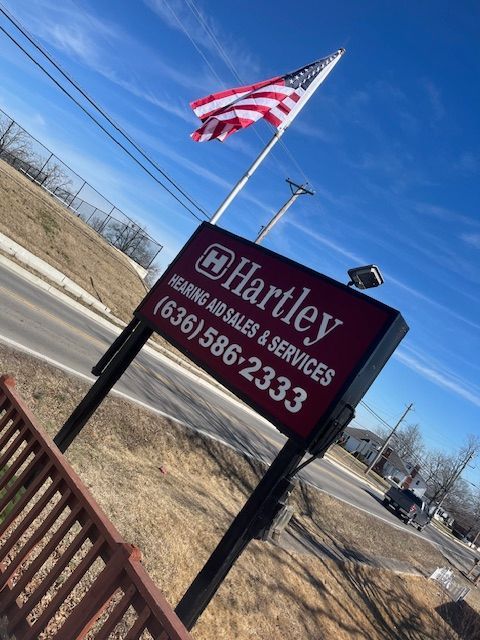Sign for Hartley Hearing Aid Sales & Services with US flag. (636)586-2333. Blue sky background.