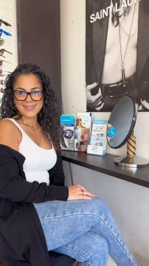 A woman wearing glasses is sitting at a desk in front of a mirror.
