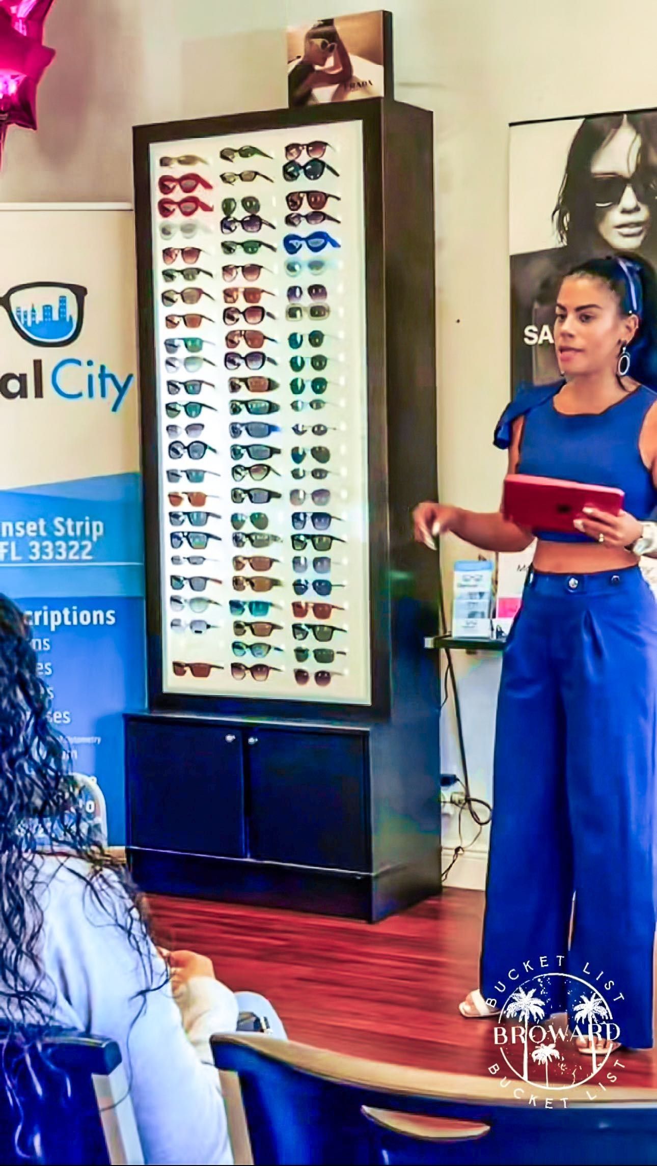A woman is standing in front of a display of glasses while holding a tablet.