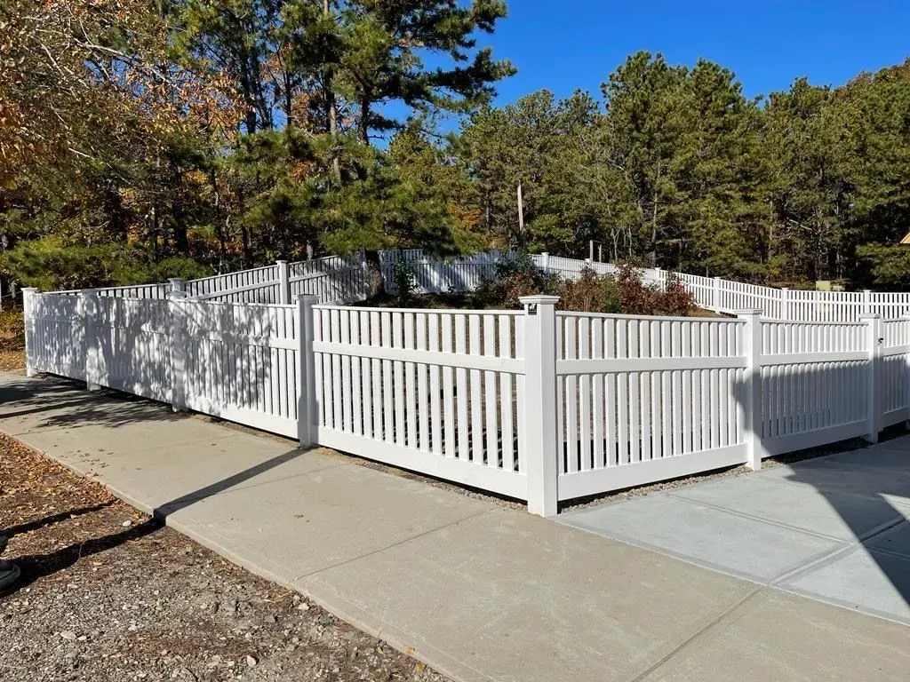White vinyl fence and gate along a concrete walkway beside landscaped trees on a sunny day.