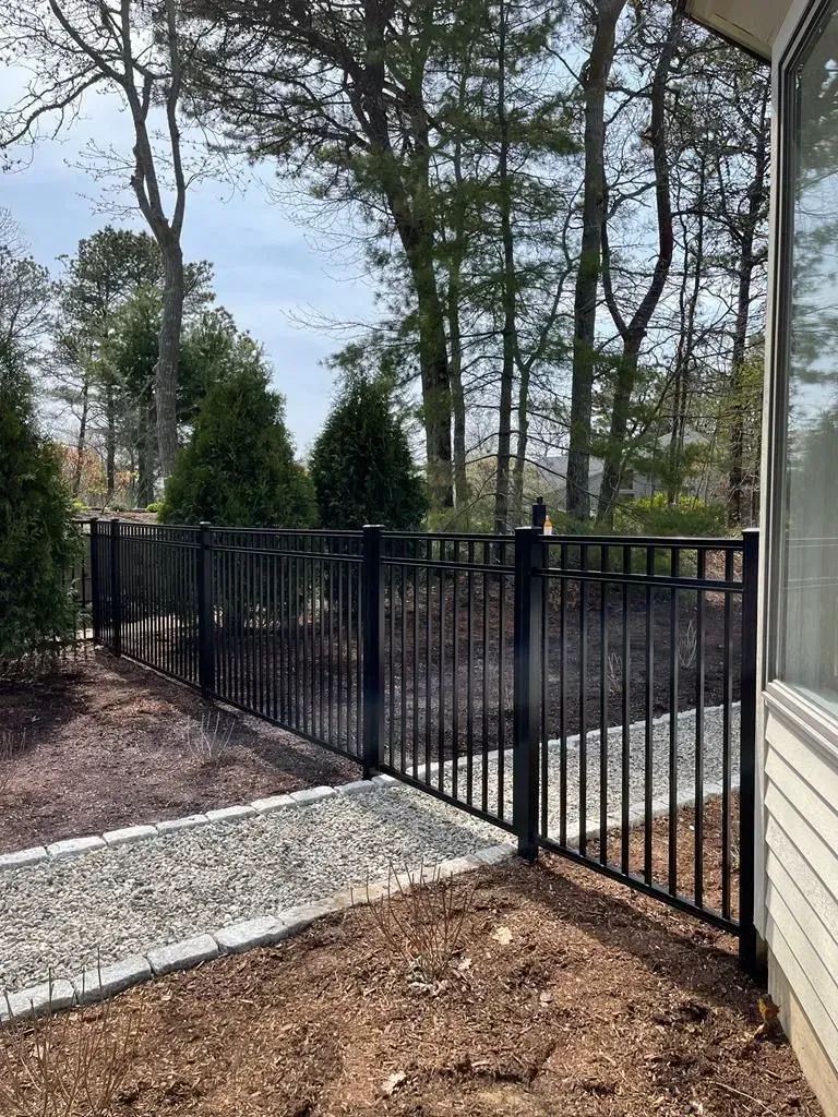 Black metal fence and gate beside a house, with gravel path and trees in the backyard.