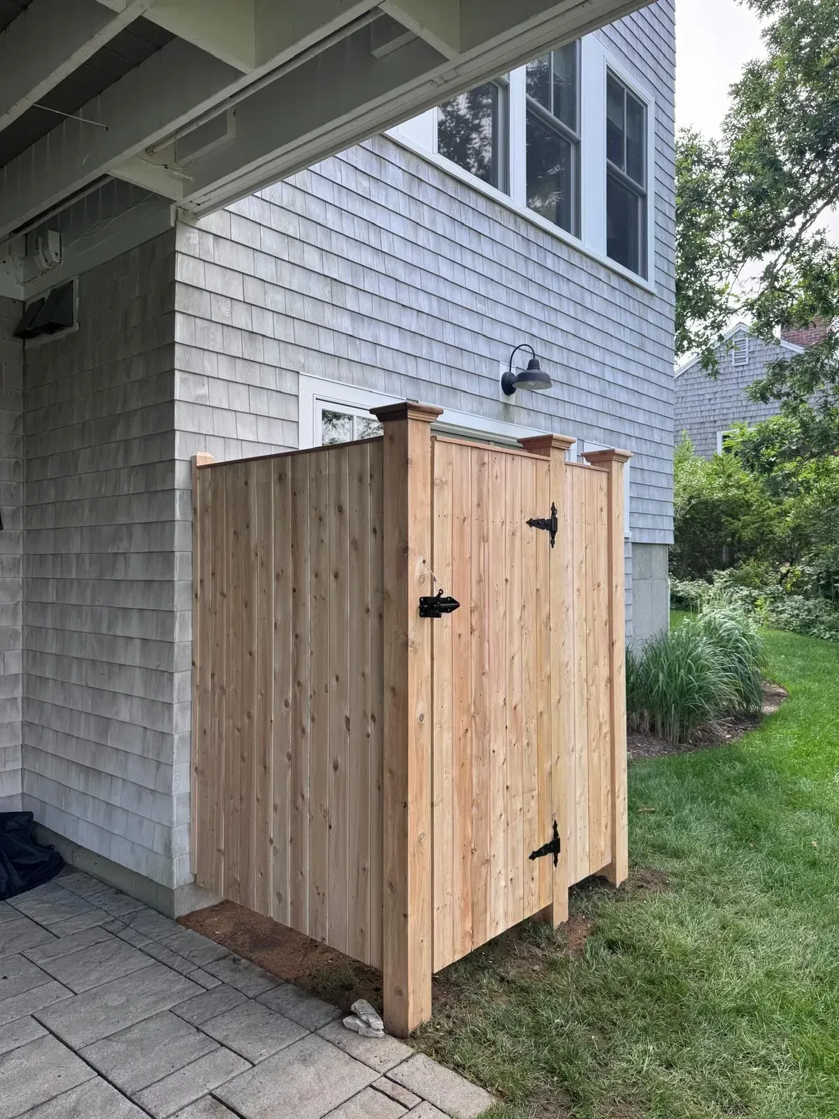 Wooden outdoor shower stall beside a house under an overhang, with black latches and a brick patio.