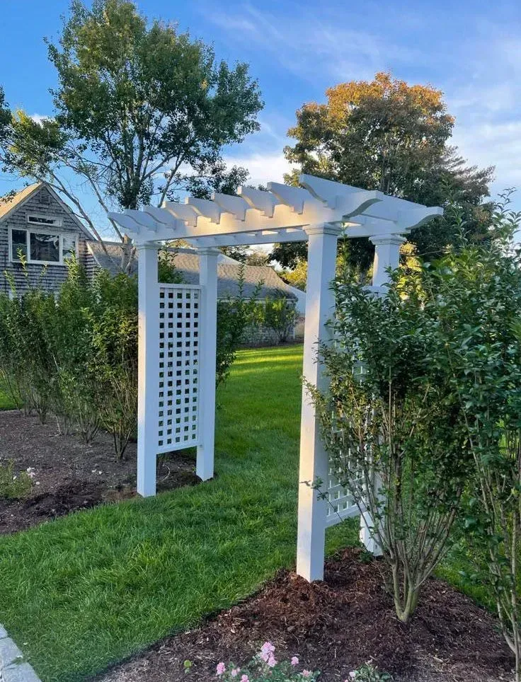 White garden arbor with trellis sides and lattice top in a landscaped backyard, surrounded by grass and shrubs.