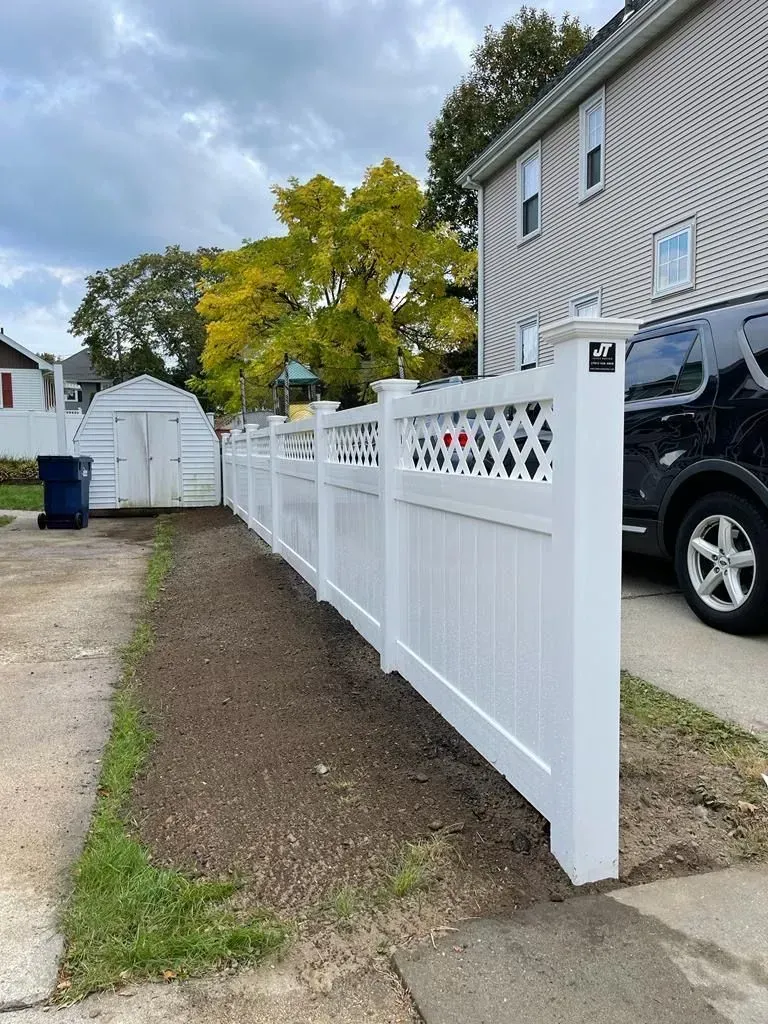 White vinyl fence along a gravel driveway beside a house and parked SUV under a cloudy sky.
