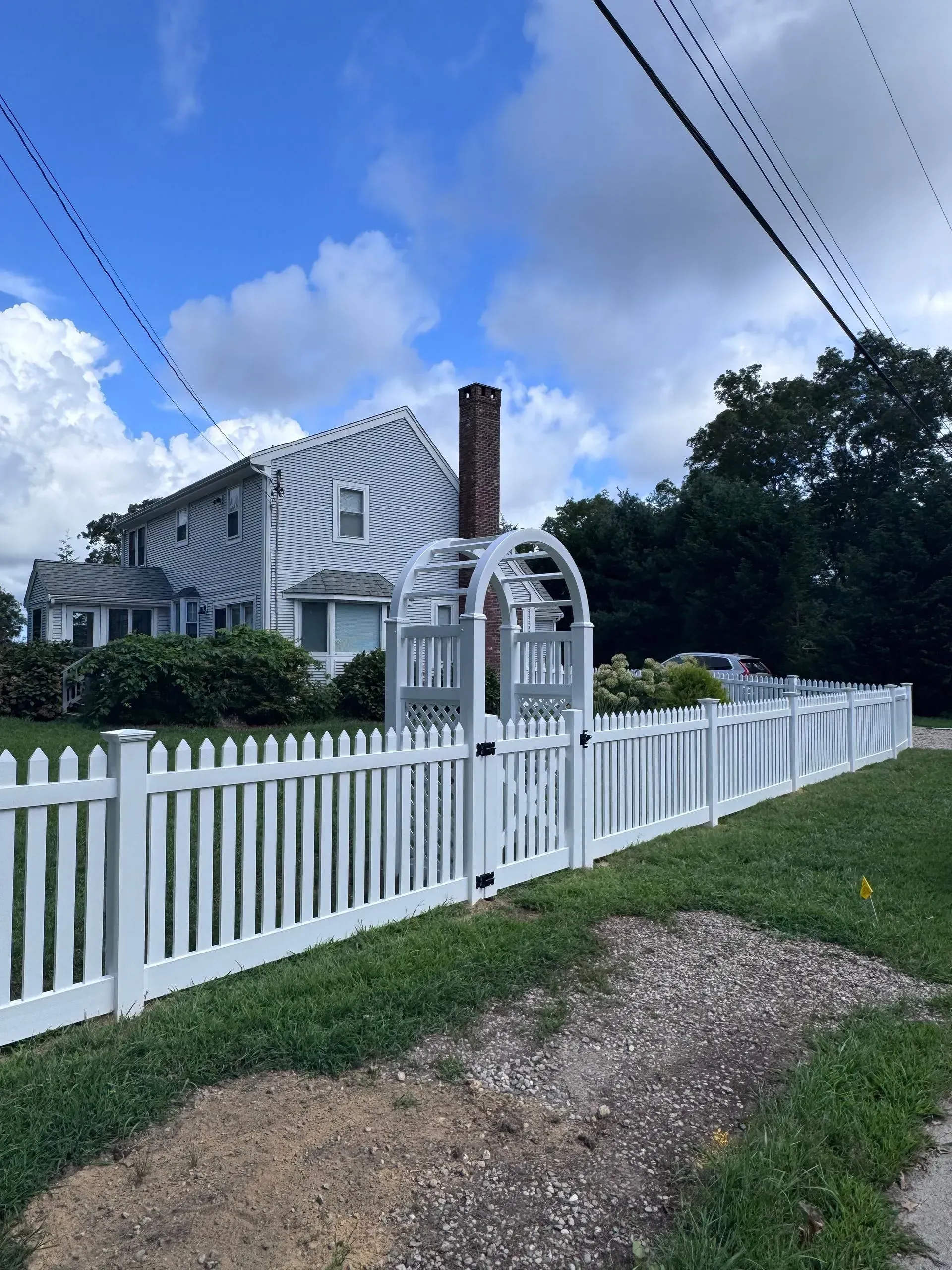 White house with chimney behind a white picket fence and gate under a blue sky with clouds.