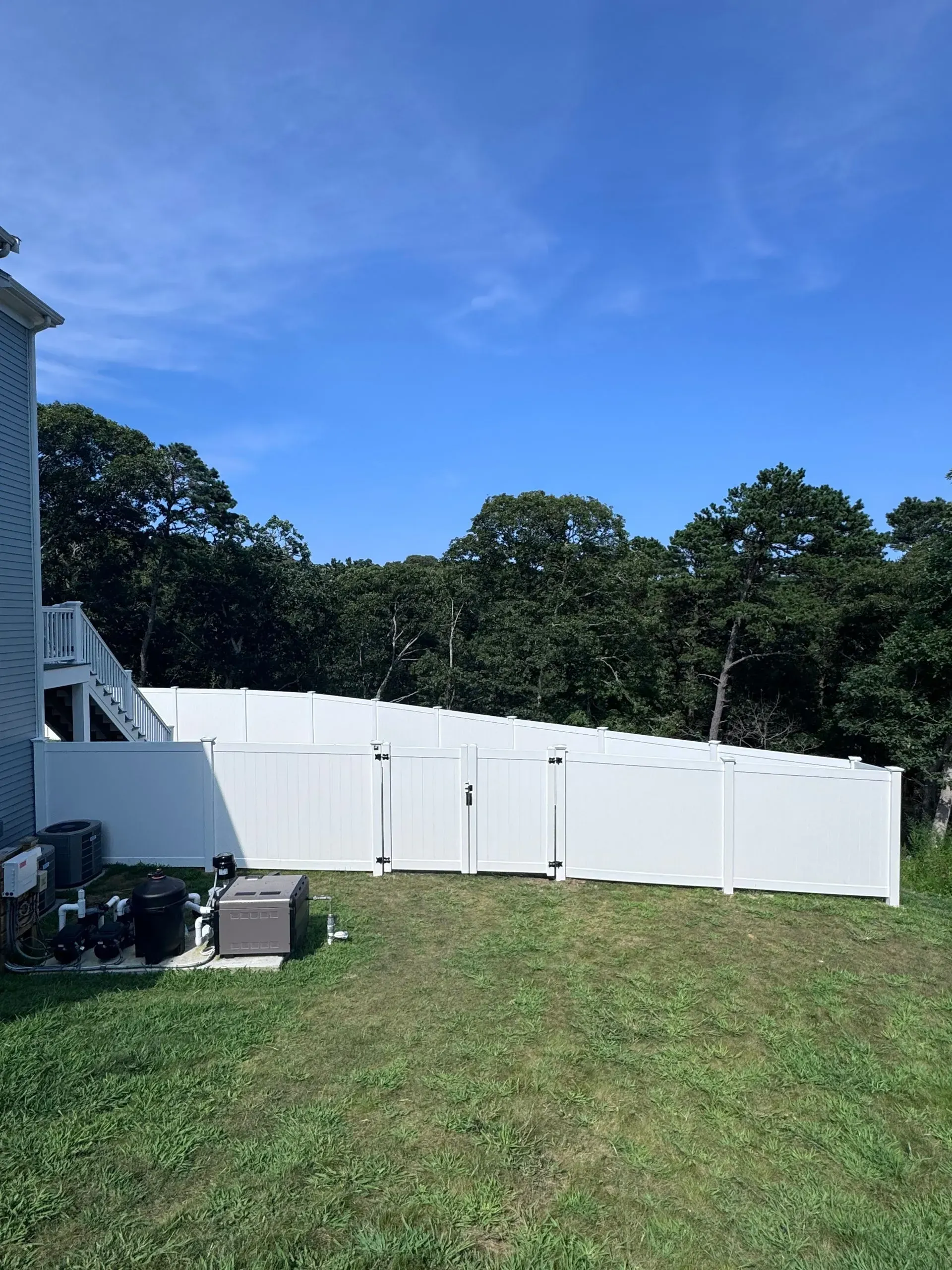 Backyard with white vinyl fence, grassy lawn, and trees under a blue sky.