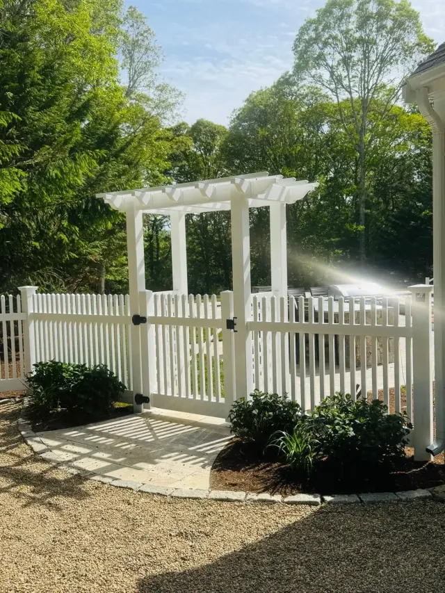 White garden gate and pergola in a fenced yard, surrounded by greenery and gravel path in sunlight.