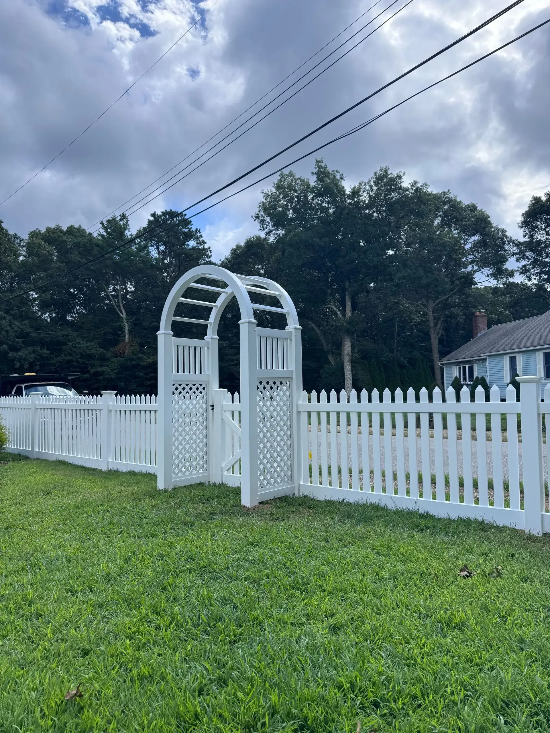 White garden gate and picket fence with archway in a grassy yard under cloudy skies.