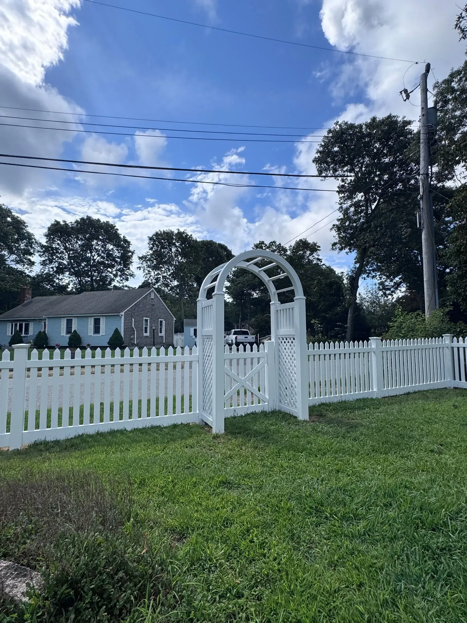 White picket fence and garden archway in a grassy yard under a cloudy blue sky.