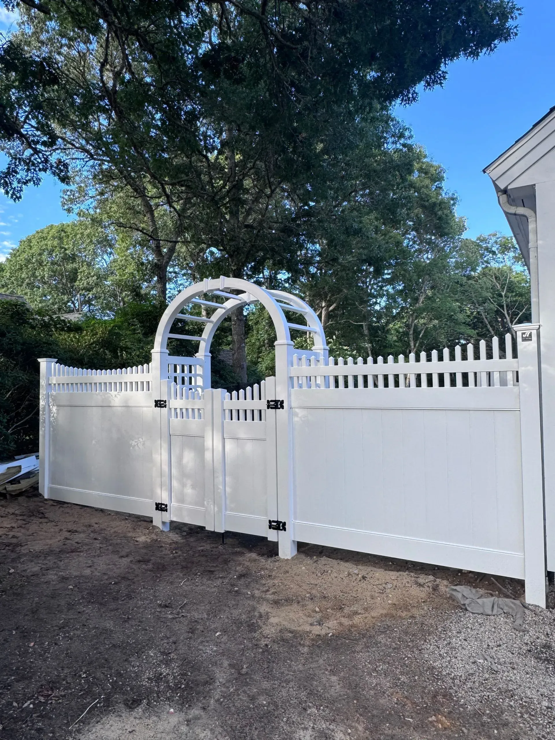 White arched garden gate and fence beside a house, with a leafy tree overhead and blue sky.