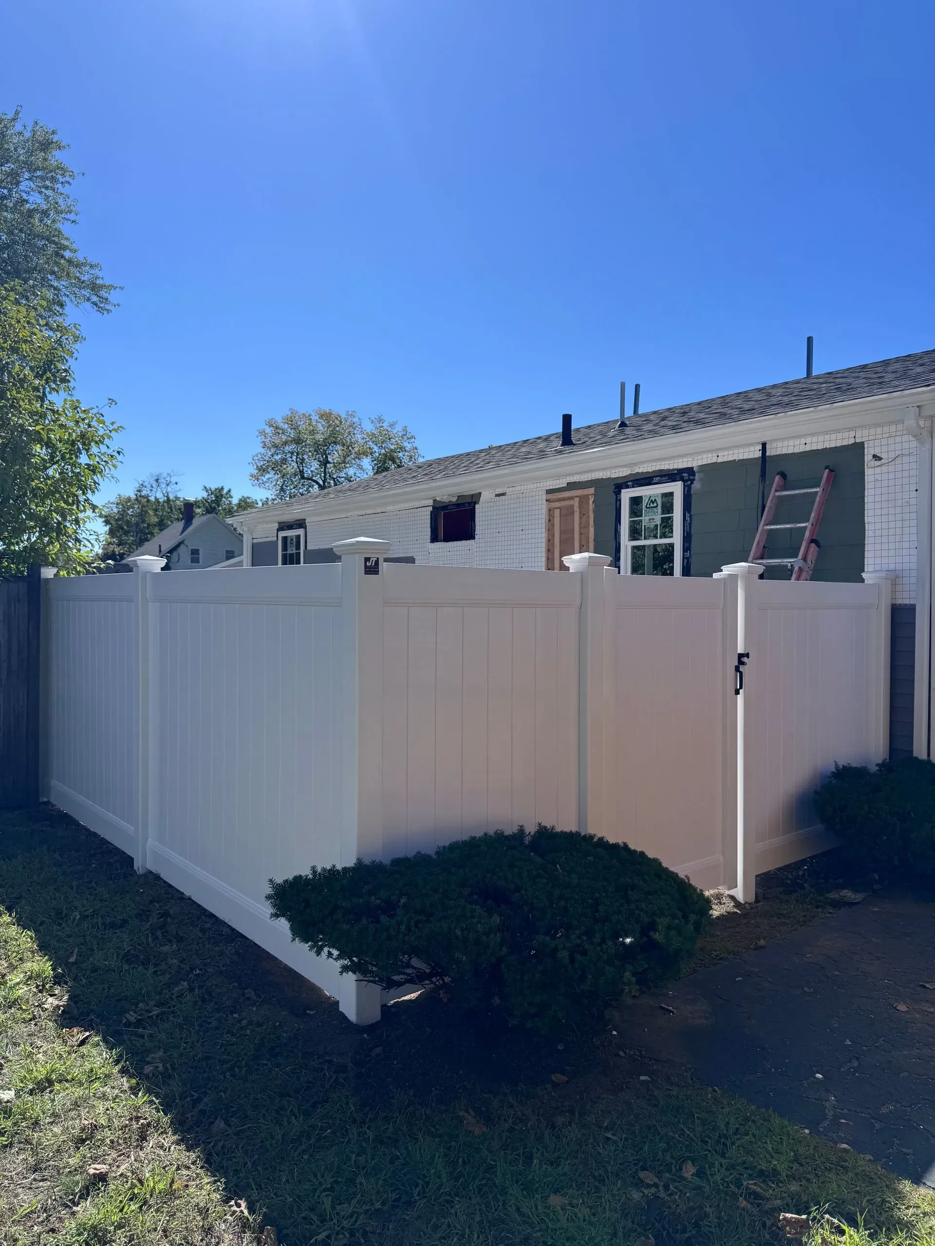 White vinyl fence around a backyard beside a beige house, with a ladder leaning against the wall.