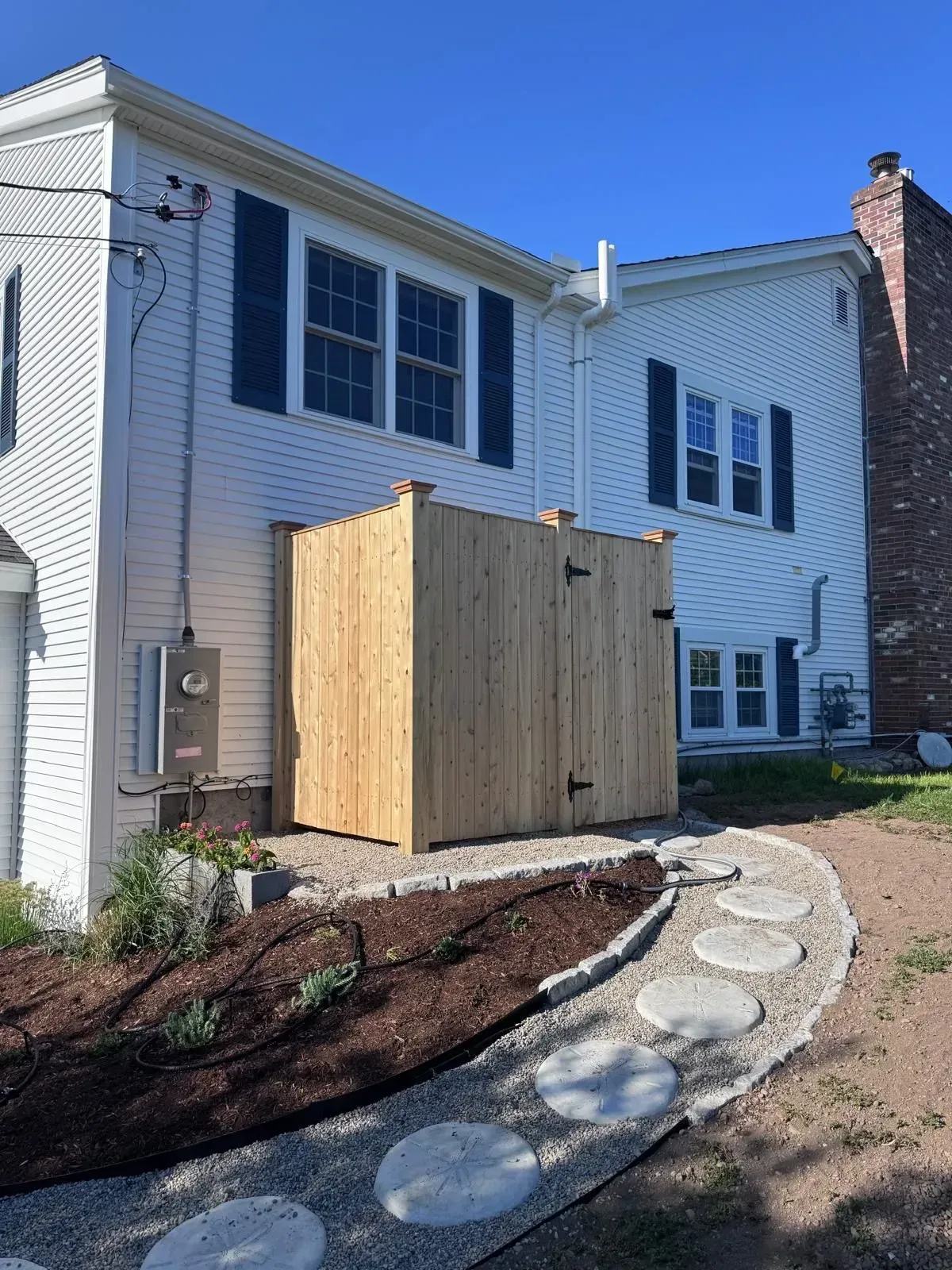 White two-story house with a wooden utility enclosure and curved stone walkway in front.