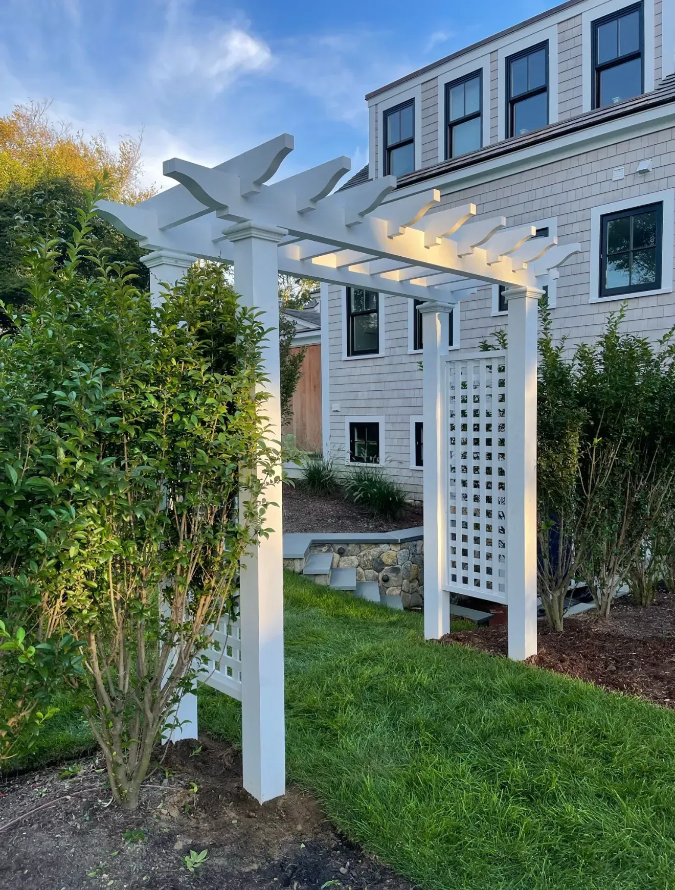 White garden pergola with bird-shaped top beside a white house and green shrubs.