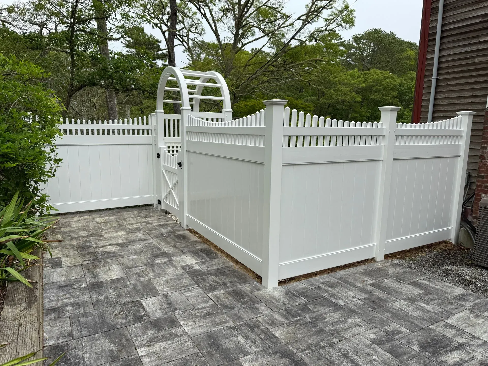 White vinyl fence and gate enclosing a paved patio beside a brick house and trees.