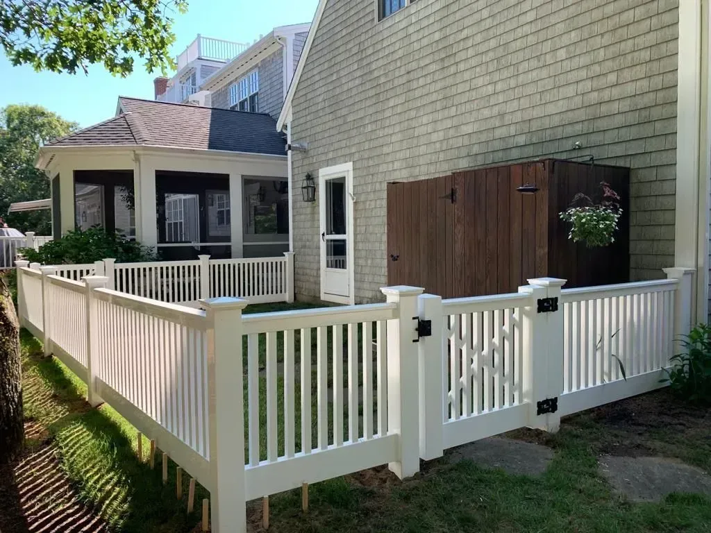White backyard fence and gate beside a beige brick house with a small porch and wooden storage paneling.