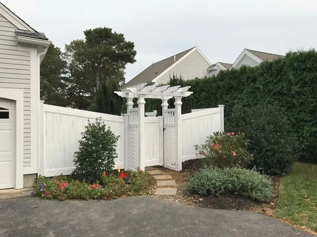 White garden arbor and gate beside a white fence, framed by shrubs and flowers in a landscaped yard.