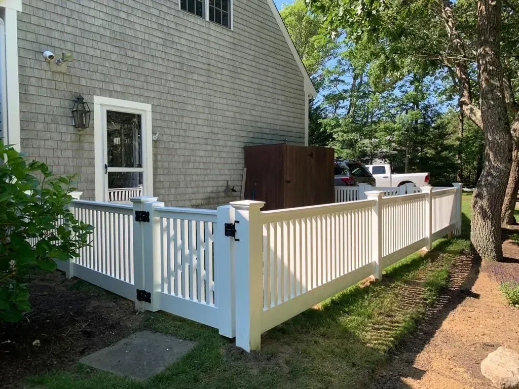 White fenced patio beside a brick house with a small gate, trees, and parked vehicles nearby.