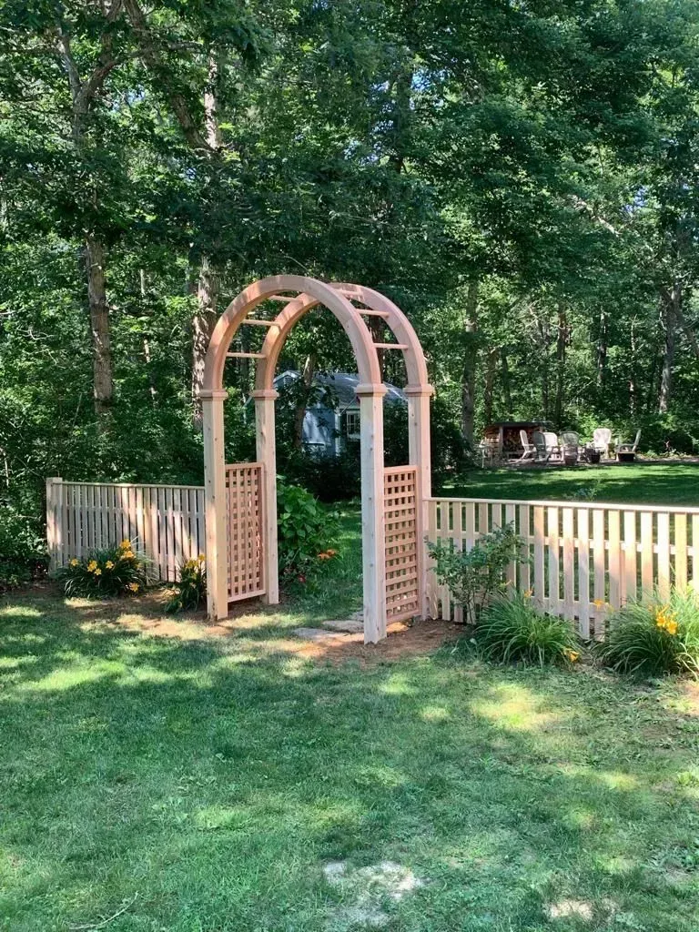 Wooden garden arbor with lattice sides in a grassy yard under trees, beside a white picket fence.