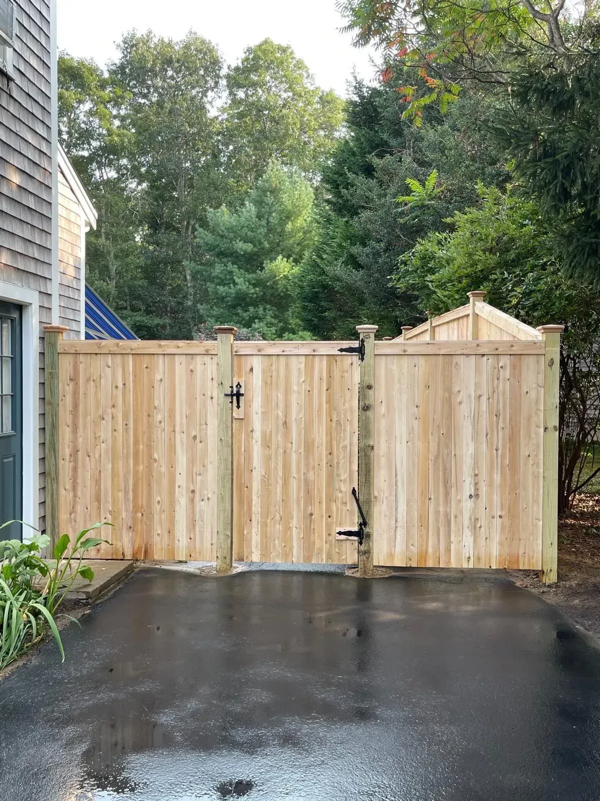 Wooden double gate at the end of a wet driveway beside a house, with trees behind it.