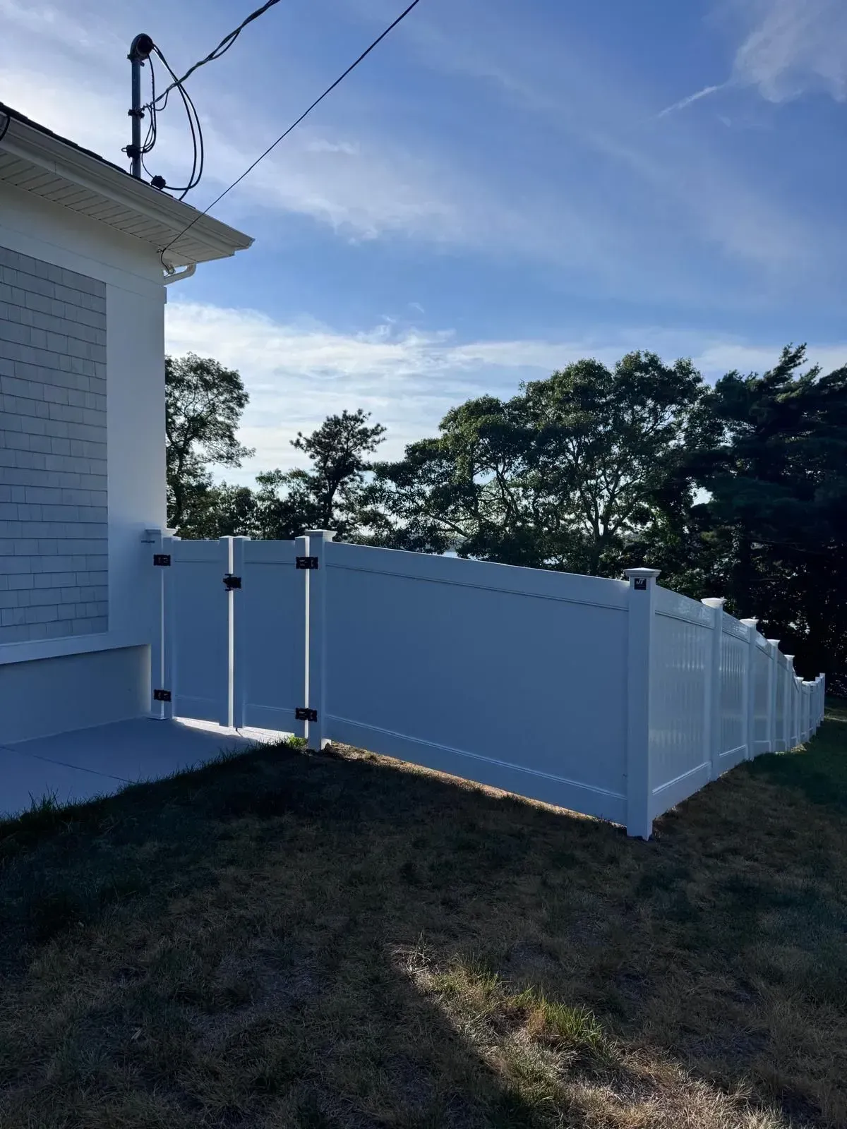 White vinyl fence beside a house on a grassy slope, with trees and blue sky in the background.