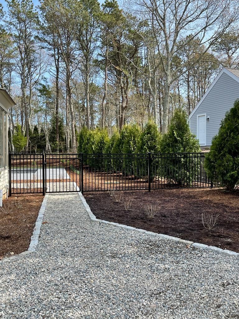 Gravel walkway leading to a black gate, with hedges, trees, and houses in the background.