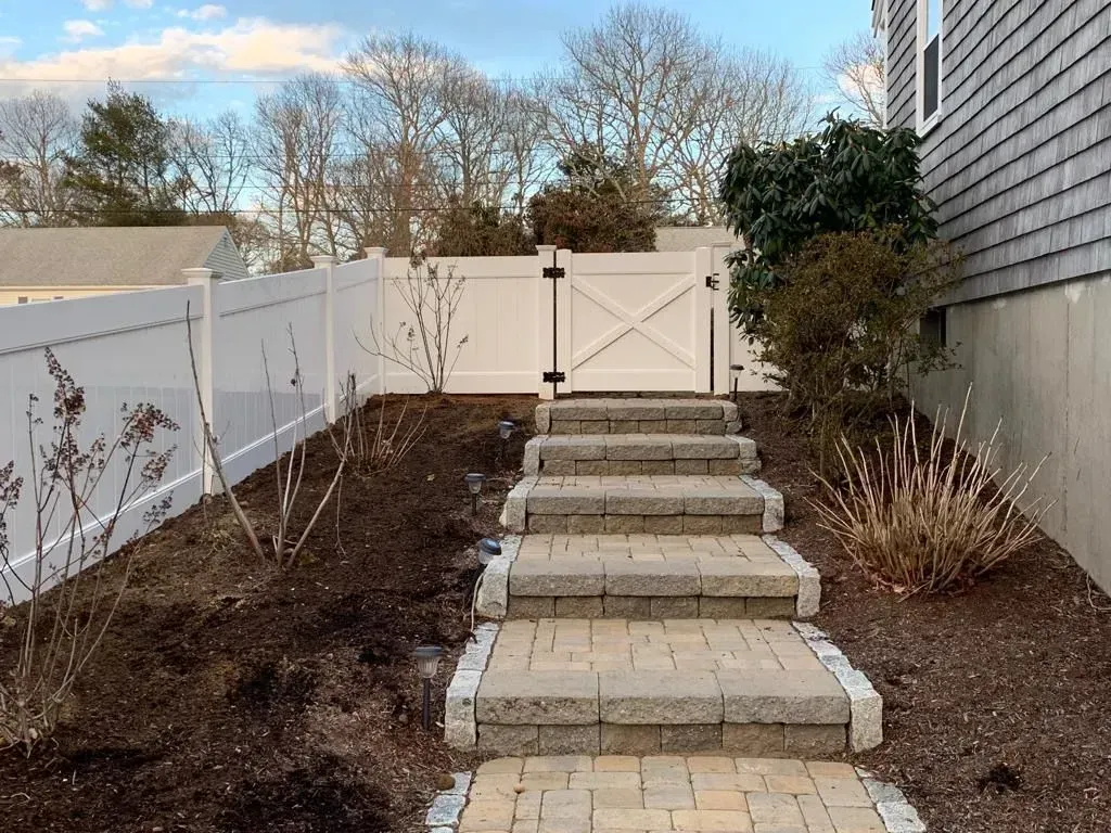 Stone steps leading to a white fence gate beside a house, with mulched landscaping and bare shrubs.